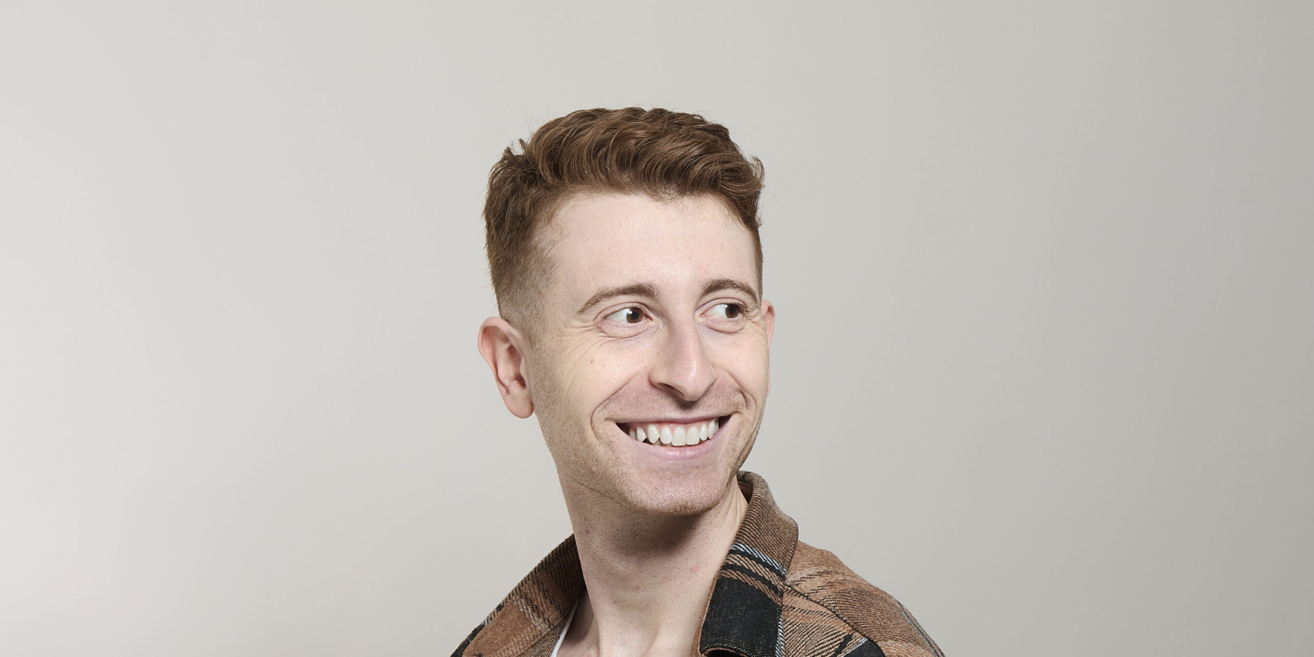 A smiling person with short, wavy hair looks to the side, wearing a patterned shirt against a neutral background.