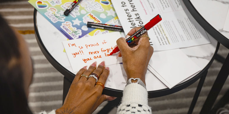 A person writes a heartfelt message on a card using a red marker, surrounded by colorful stickers and papers.