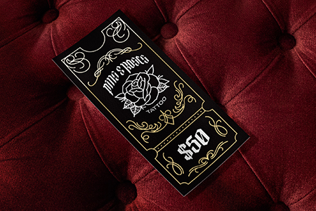 A black tattoo gift certificate with a rose design, valued at $50, rests on a red velvet surface.
