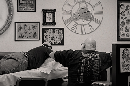 A tattoo artist works on a client lying on a table in a tattoo studio, surrounded by artwork and a clock.