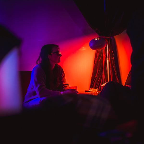 A woman sits in a dimly lit room, holding a drink, with colorful lighting and a lamp nearby.