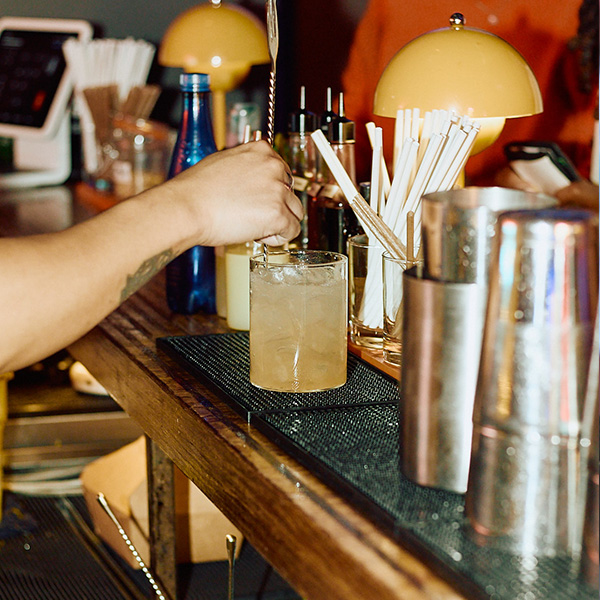 A hand stirs a cocktail in a glass at a bar, surrounded by bottles and bar tools.