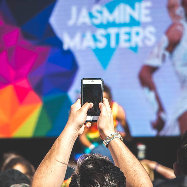 A person holds a smartphone to capture a performance on stage, with colorful background and text "Jasmine Masters.