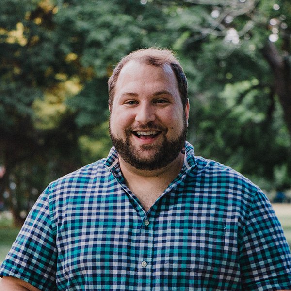 A smiling man with a beard wears a blue and green plaid shirt in a park setting.