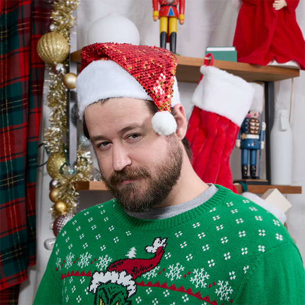 A man with a beard wears a festive red and white hat and a green Christmas sweater, posing in a decorated room.