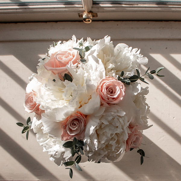A round bouquet of white peonies and pink roses, accented with green leaves, rests on a windowsill.