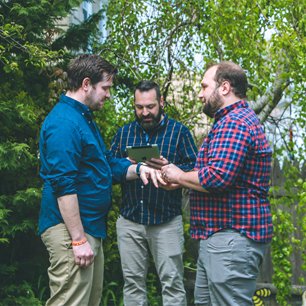 Three men stand outdoors, one holding a tablet while examining another's wrist.