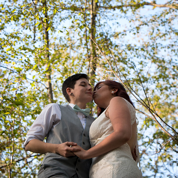Two brides share a kiss in a forest, surrounded by greenery, celebrating their wedding.