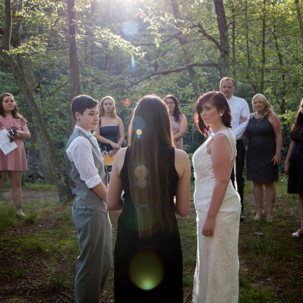 Two people in wedding attire stand facing each other, surrounded by guests in a wooded setting.