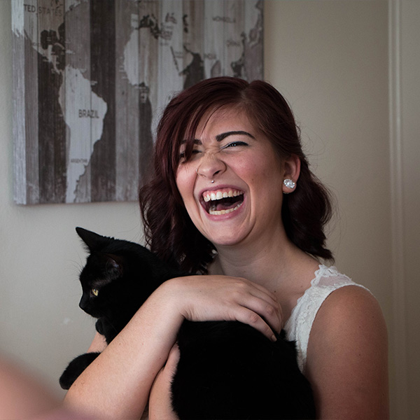 A woman in a wedding dress laughs while holding a black cat. A world map hangs in the background.