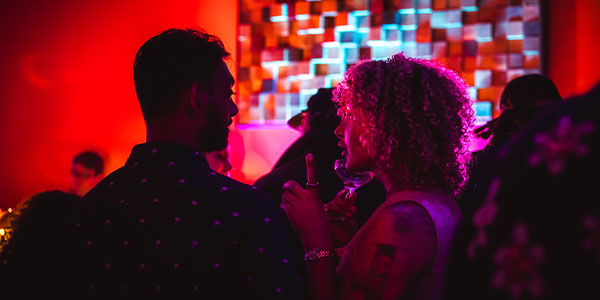 Two people engage in conversation at a lively event, colorful lights in the background.