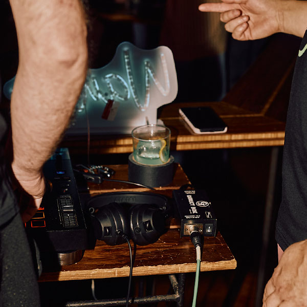 Two people interact at a DJ setup with headphones, a drink, and a glowing sign in the background.
