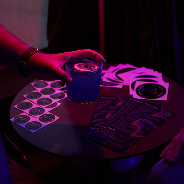 A hand holds a blue cocktail near a table with menus and coasters in dim lighting.